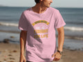 A man proudly wears a pink Garment Graphics Fishing T-shirt on the beach, featuring Father and Son: Fishing Partners For Life in yellow text. The design includes a fish and fishing rod, symbolizing family bonding.