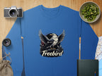 A Garment Graphics sweatshirt with an eagle design is placed on a wooden surface alongside a camera, lens, bowl of kiwis, black notebook, map, and burlap bag. The unisex sweatshirt features a bird in flight illustration with the word Freebird.