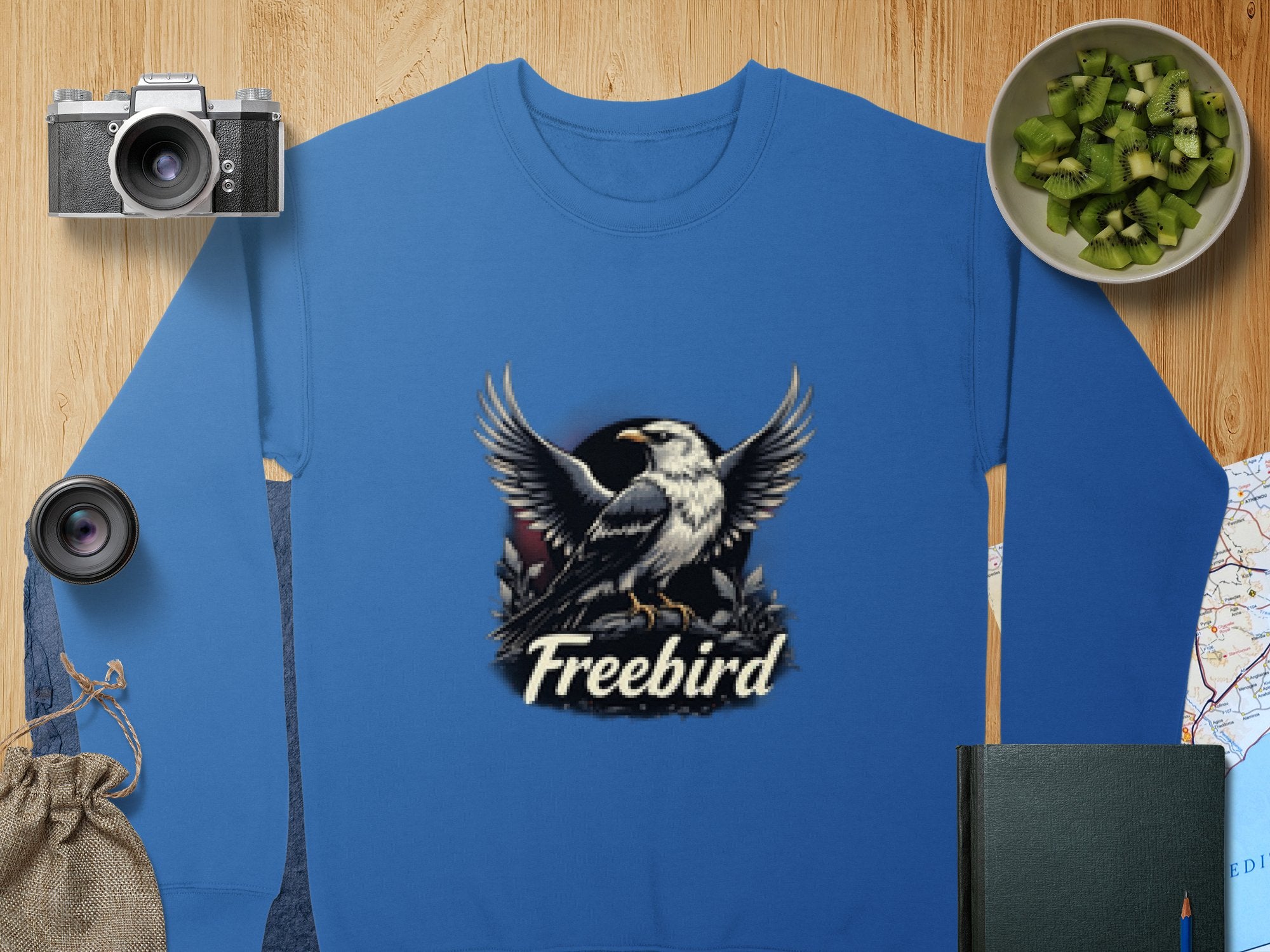 A Garment Graphics sweatshirt with an eagle design is placed on a wooden surface alongside a camera, lens, bowl of kiwis, black notebook, map, and burlap bag. The unisex sweatshirt features a bird in flight illustration with the word Freebird.