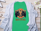 A green Garment Graphics t-shirt with two bull heads above BULLOCKS, part of the Bullocks Pintable collection, is on a wooden surface with a light gray cardigan. White flowers and small items surround it.