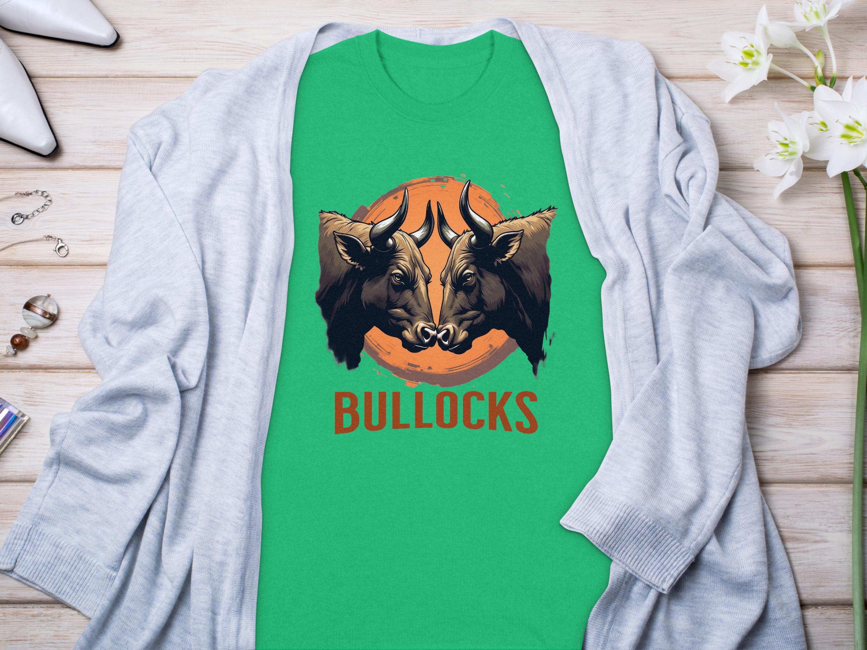 A green Garment Graphics t-shirt with two bull heads above BULLOCKS, part of the Bullocks Pintable collection, is on a wooden surface with a light gray cardigan. White flowers and small items surround it.