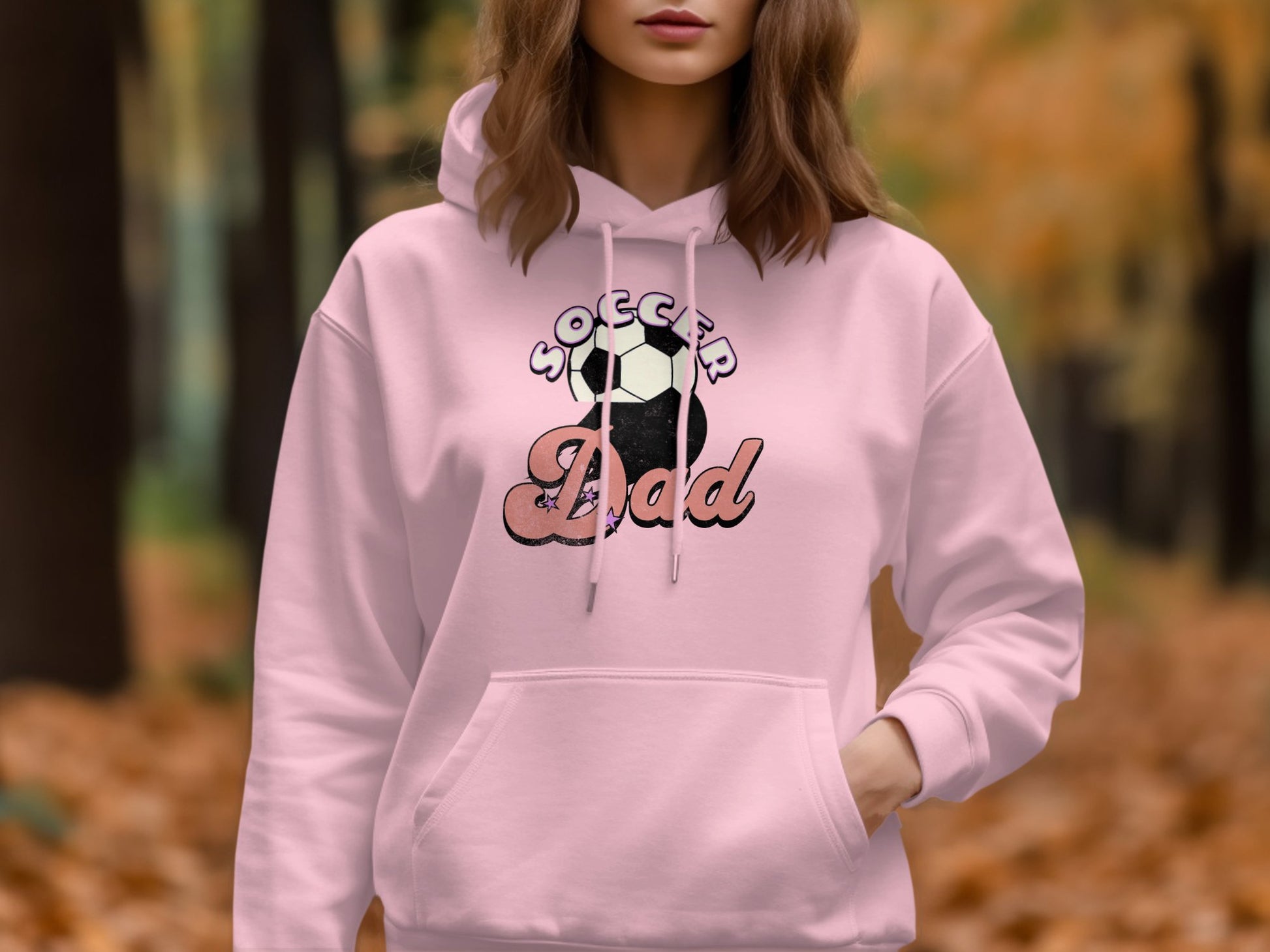 In a forest filled with autumn leaves, someone wears a pink Soccer Dad hoodie by Garment Graphics. The soccer ball graphic stands out against the blurred fall setting, merging casual style with the seasons vibrant essence.