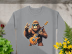 Garment Graphics gray sweatshirt showcases a gorilla in sunglasses, rocking an electric guitar and a peace sign. Its placed on concrete with green leaves and yellow flowers, ideal for music enthusiasts.
