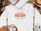 Garment Graphics white hoodie features a youre my cup of tea design in brown, displayed alongside a straw hat, sunglasses, spiral notebook, map, brown sandals, woven bag, and white camera on wood—a perfect setup for capturing cozy moments.