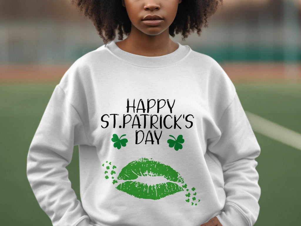 A person is wearing a Garment Graphics white sweatshirt with Happy St. Patricks Day in black text, two green clovers, a large green lip print symbolizing a clover kiss, and small stars. The blurred background adds to the festive spirit.