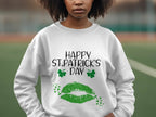 A person is wearing a Garment Graphics white sweatshirt with Happy St. Patricks Day in black text, two green clovers, a large green lip print symbolizing a clover kiss, and small stars. The blurred background adds to the festive spirit.