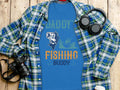 Garment Graphics Daddys Fishing Buddy Graphic Print T-Shirt is adorned with fishing graphics, ideal for enthusiasts. Styled on wood with a plaid shirt, its surrounded by a camera, headphones, and a white travel mug for an artistic touch.