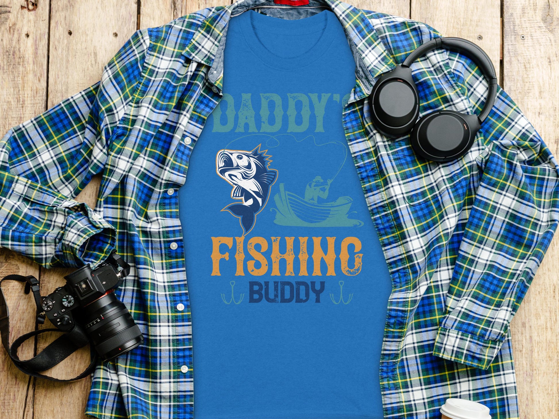 Garment Graphics Daddys Fishing Buddy Graphic Print T-Shirt is adorned with fishing graphics, ideal for enthusiasts. Styled on wood with a plaid shirt, its surrounded by a camera, headphones, and a white travel mug for an artistic touch.