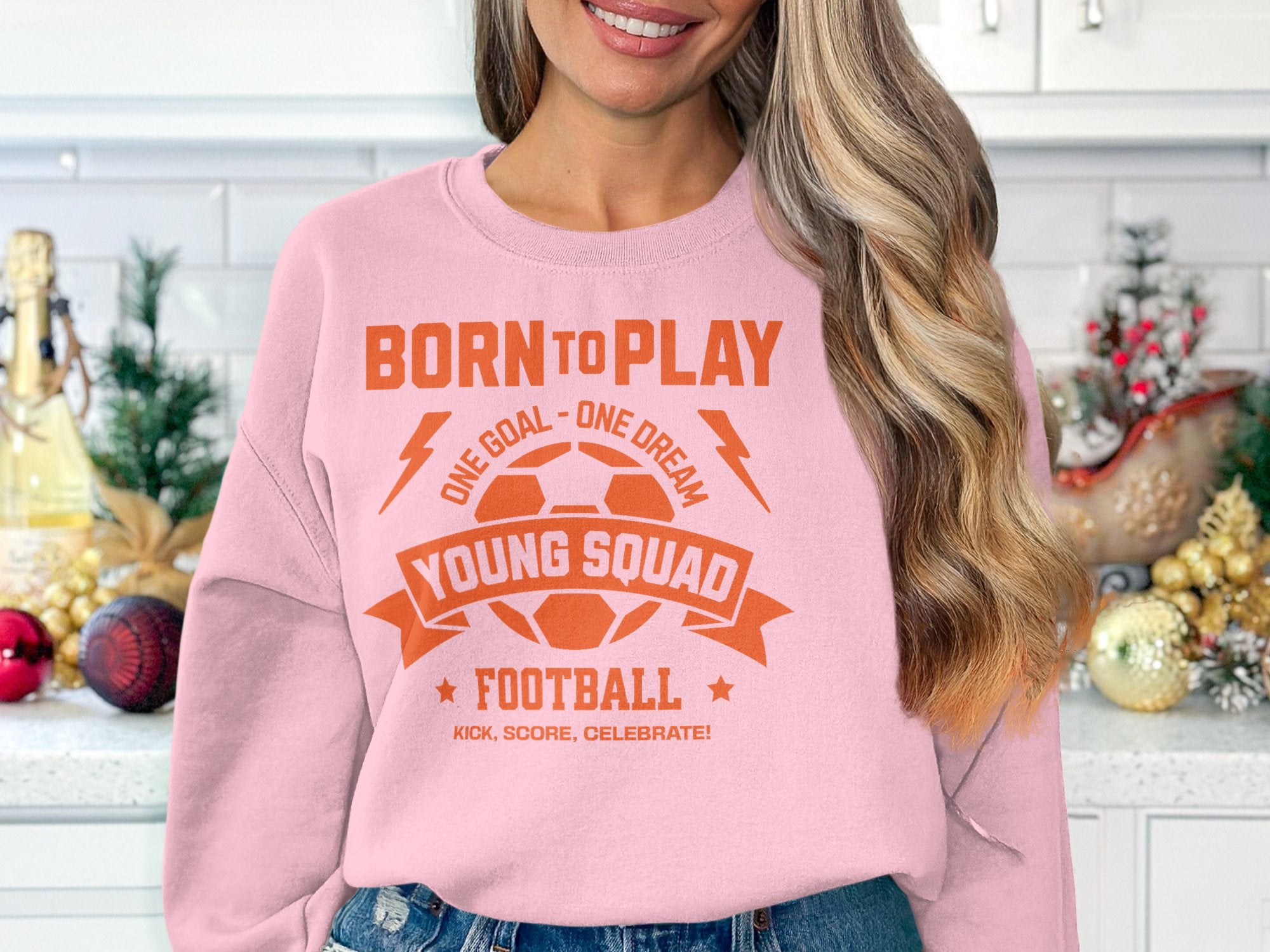 A woman with long, wavy hair wears a pink Garment Graphics football sweatshirt with Born to Play, One Goal - One Dream, Young Squads! in orange. She stands in a festively decorated kitchen. Its unisex sizing makes it an ideal gift for any fan this holiday season.