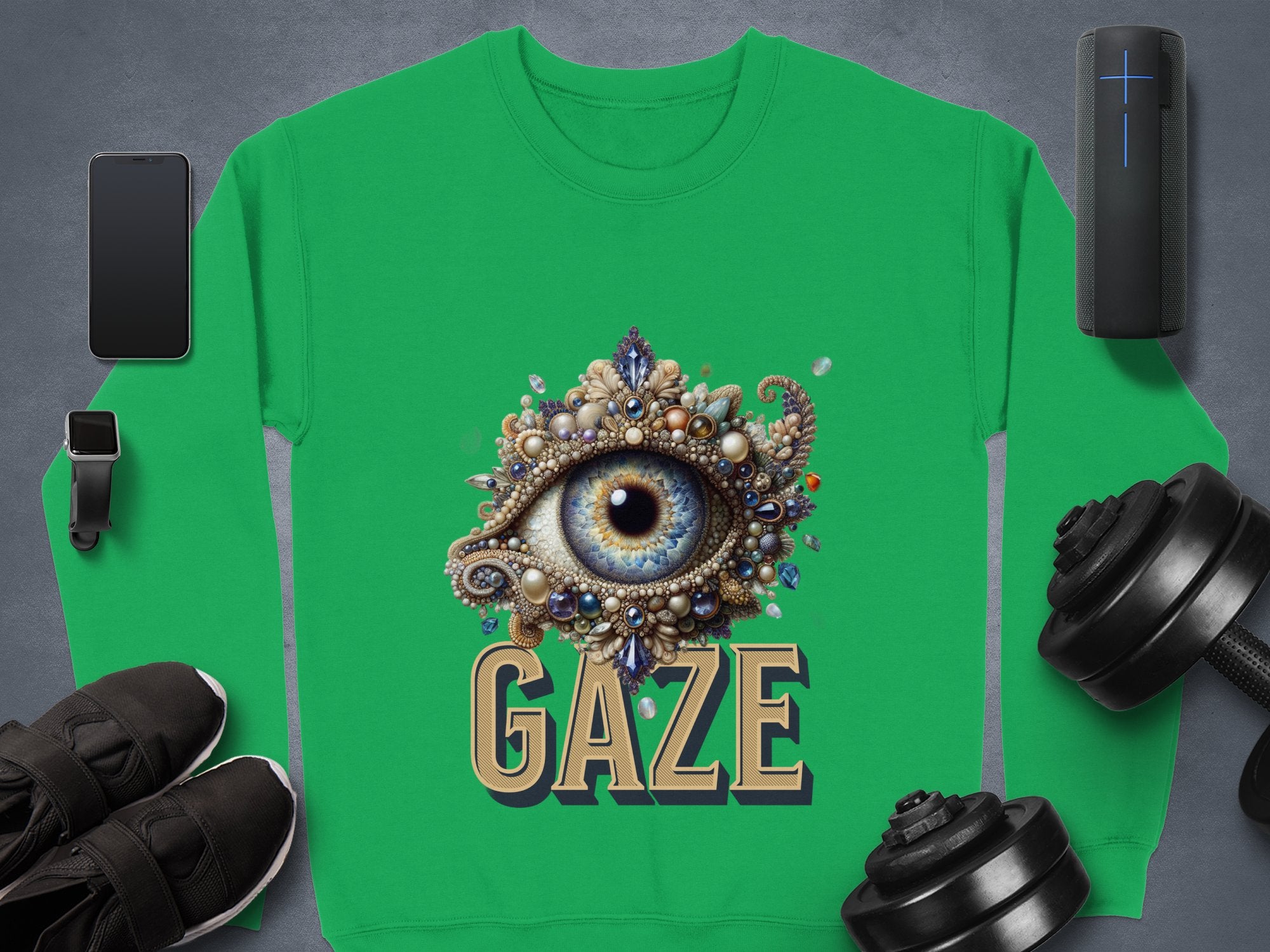 The Garment Graphics unisex graphic sweatshirt showcases an ornate eye design above GAZE, surrounded by a smartphone, smartwatch, wireless speaker, black shoes, and dumbbells on a gray background.