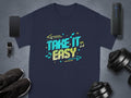 Navy blue T-shirt by Garment Graphics with vibrant Take It Easy design in colorful letters, surrounded by musical notes and arrows, laid on a gray surface near a smartphone, smartwatch, sneakers, speaker, and dumbbell.