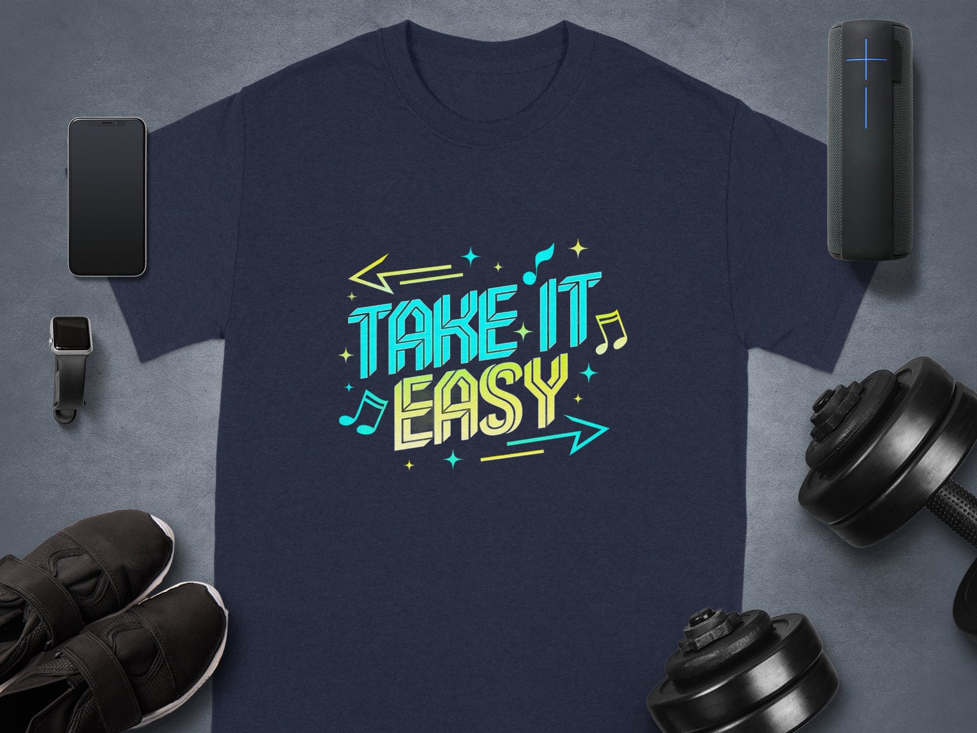 Navy blue T-shirt by Garment Graphics with vibrant Take It Easy design in colorful letters, surrounded by musical notes and arrows, laid on a gray surface near a smartphone, smartwatch, sneakers, speaker, and dumbbell.