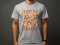 Someone dons a dark gray Garment Graphics Rodeo T-Shirt, featuring a vintage-style cowboy illustration. Enthusiasts will love the brown text Howdy Rodeo and Notorious set against the background.