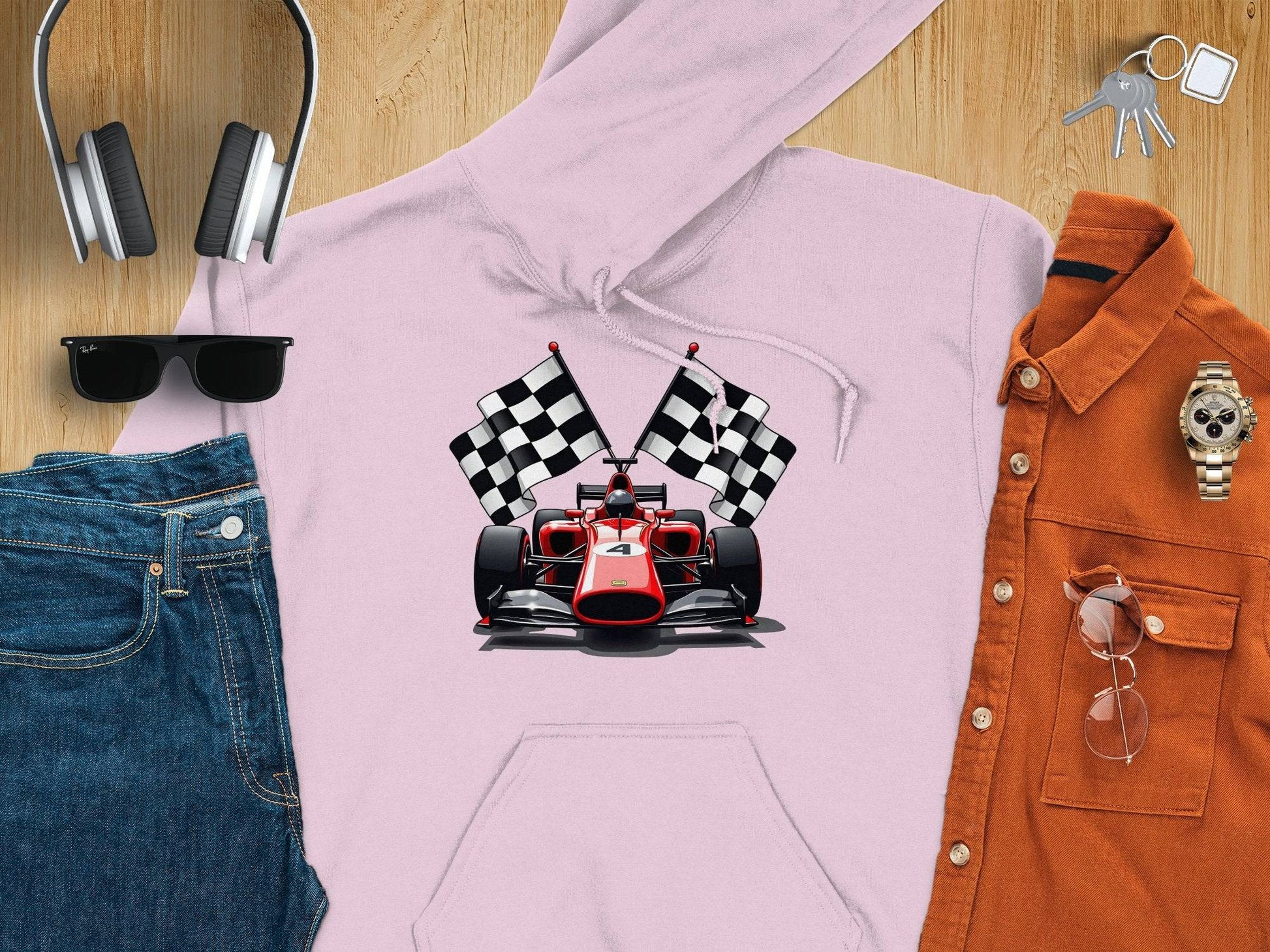 A Garment Graphics pink hoodie with a bold red racing car and two checkered flags is on a wooden surface, surrounded by headphones, sunglasses, blue jeans, an orange shirt, a watch, keys, and a smartphone.