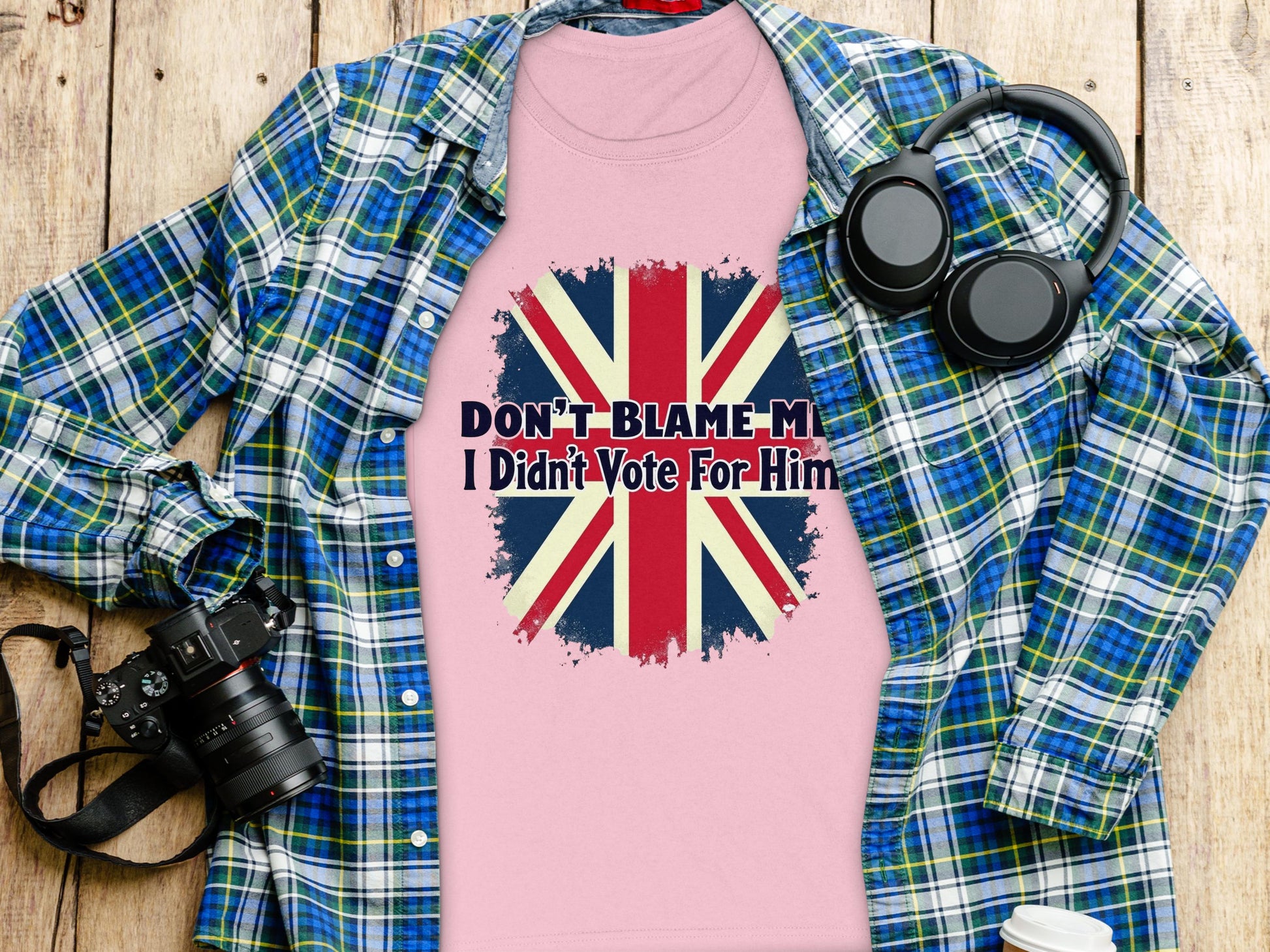 Garment Graphics presents a pink 100% cotton T-shirt featuring a distressed Union Jack and the text Dont Blame Me, I Didnt Vote For Him, artfully displayed on wood with a blue plaid shirt, headphones, camera, and white cup.