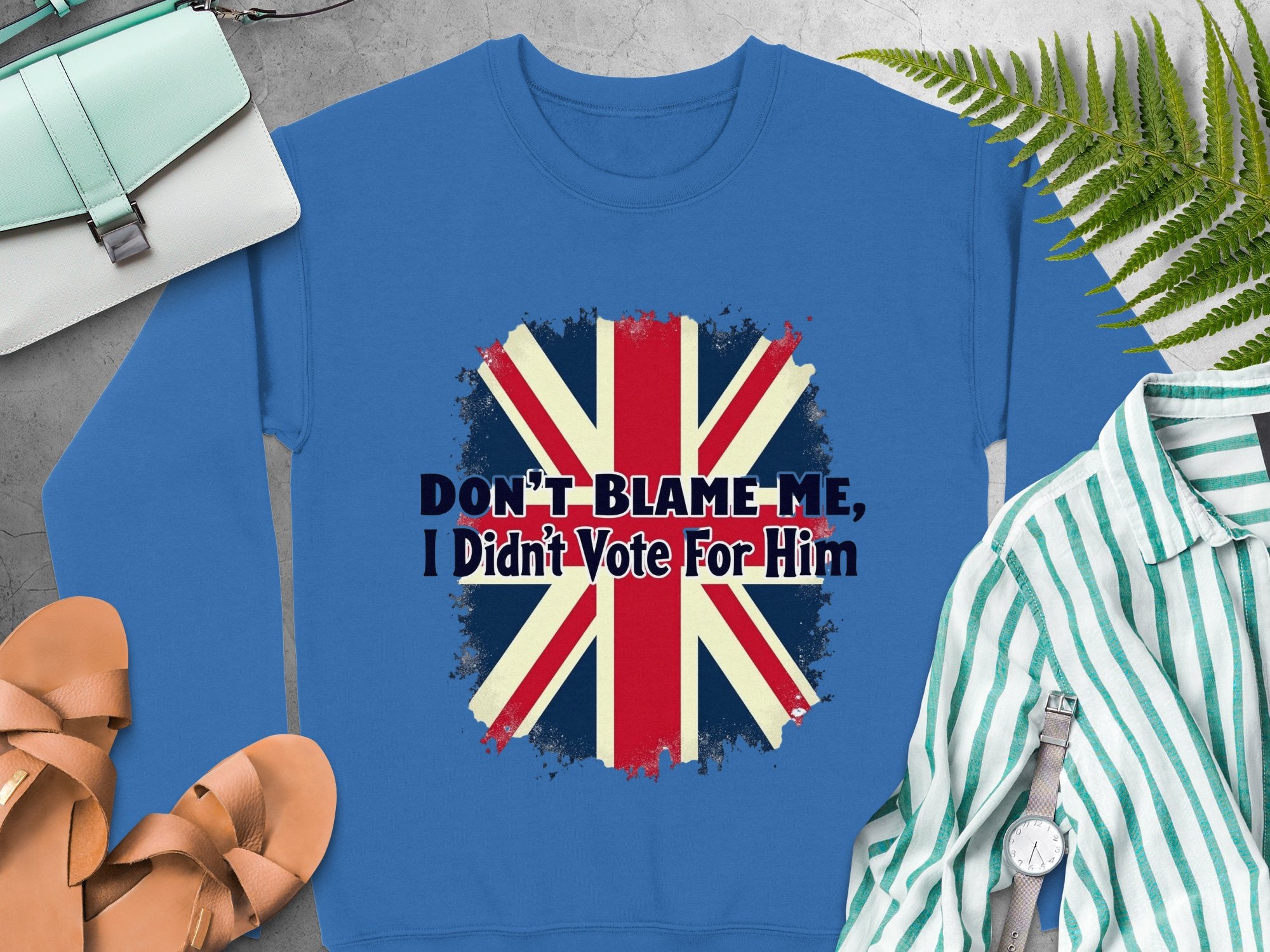 A blue Union Jack sweatshirt with Dont Blame Me, I Didnt Vote For Him from Garment Graphics offers unisex sizing and is styled with sandals, a green striped shirt, a light blue purse, and fern leaves.