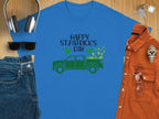 Flat lay of Garment Graphics blue T-shirt with Happy St. Patrick’s Day and a green truck with shamrocks. Includes sunglasses, denim jeans, orange shirt, watch, glasses, and headphones on wood surface, celebrating Irish pride.