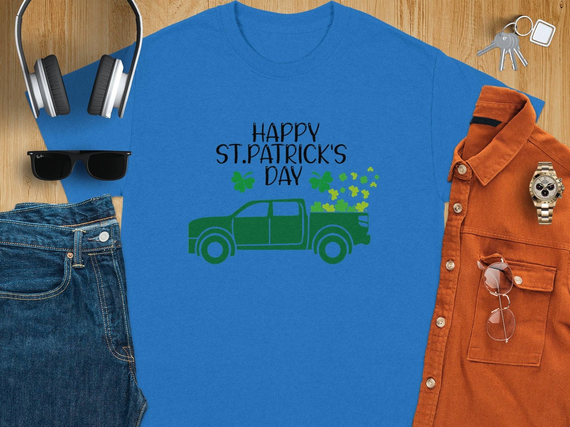 Flat lay of Garment Graphics blue T-shirt with Happy St. Patrick’s Day and a green truck with shamrocks. Includes sunglasses, denim jeans, orange shirt, watch, glasses, and headphones on wood surface, celebrating Irish pride.