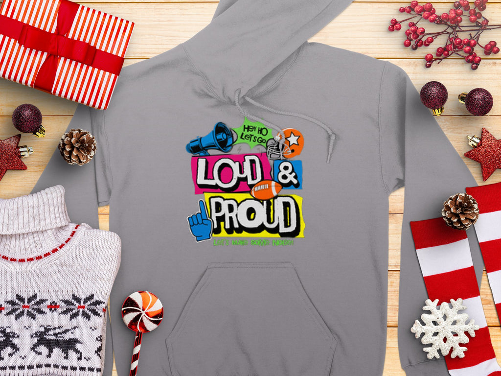 Garment Graphics Lets Make Some Noise Hoodie, with its colorful megaphone, star, whistle, and foam finger graphic reading LOUD & PROUD and Spirit Wear Your Way, lies on a wooden surface amidst festive decorations like gifts, candy, and pinecones.