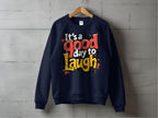 A navy blue, unisex sweatshirt from Garment Graphics hangs on a wooden hanger against a concrete wall. It features bold, colorful text reading Its a Good Day to Laugh.