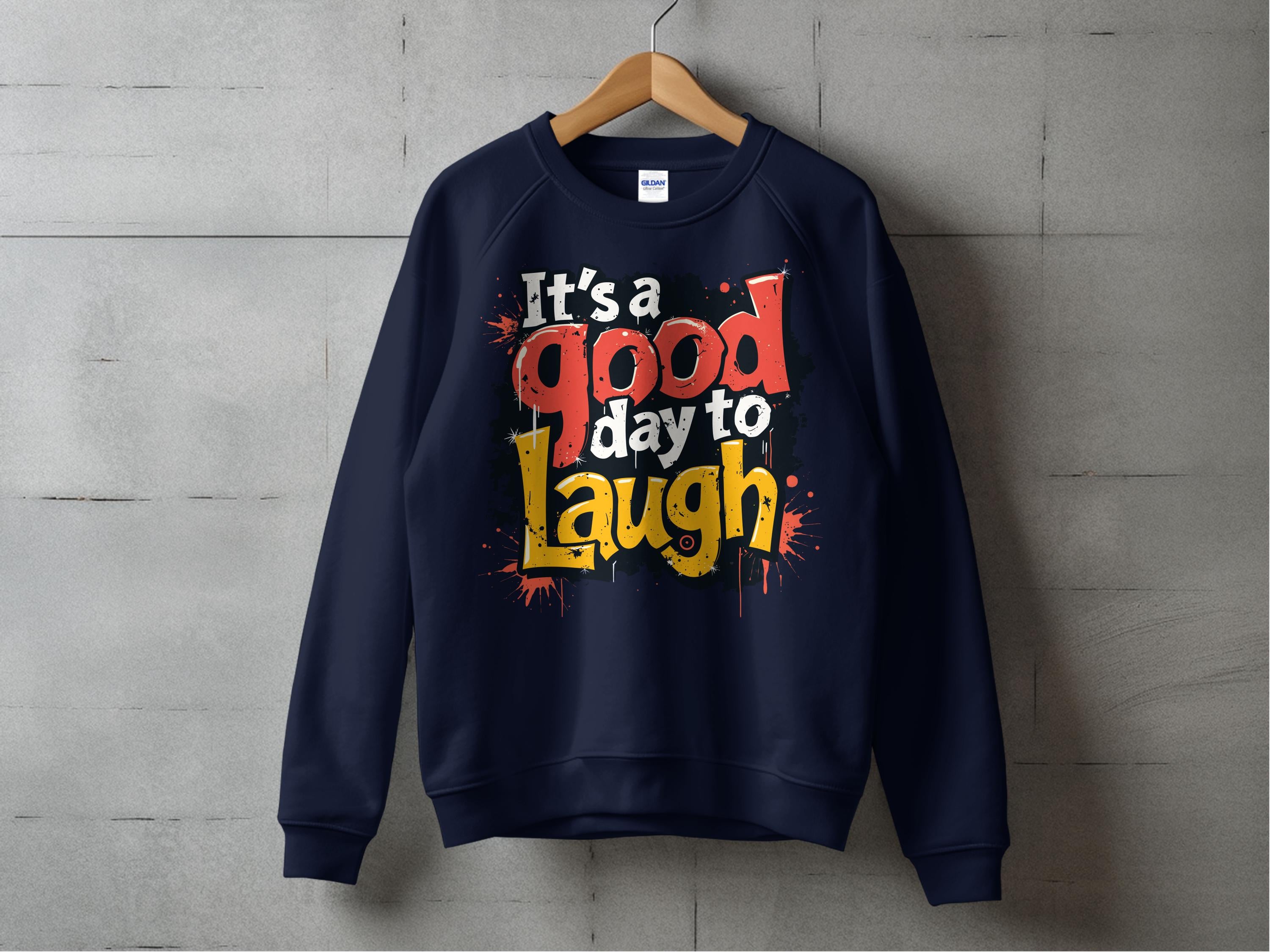 A navy blue, unisex sweatshirt from Garment Graphics hangs on a wooden hanger against a concrete wall. It features bold, colorful text reading Its a Good Day to Laugh.