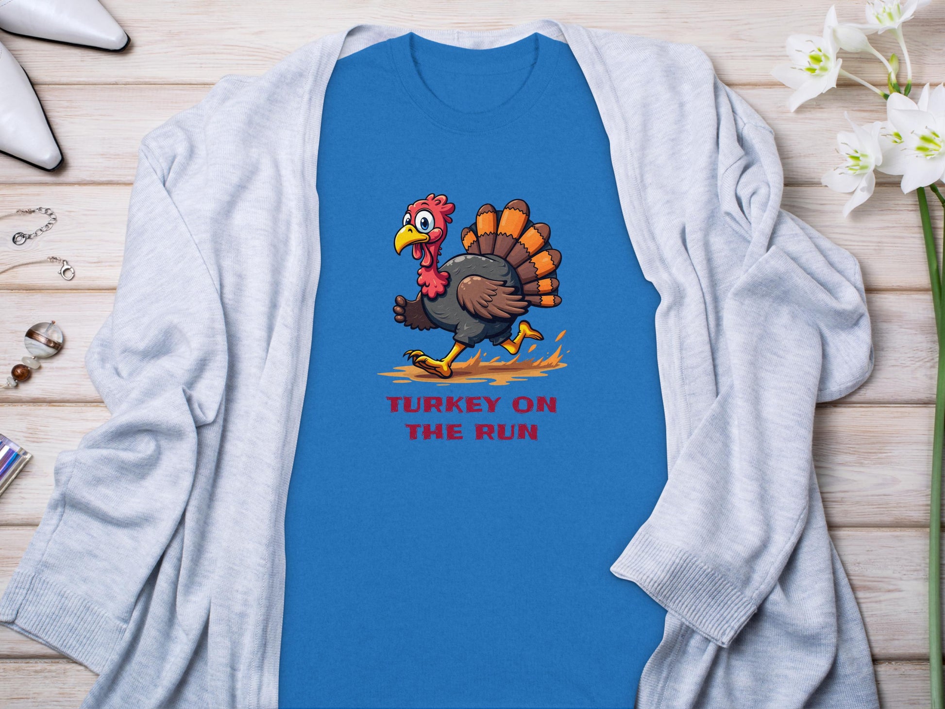 Garment Graphics blue T-shirt features a cartoon turkey in yellow shoes with TURKEY ON THE RUN in red below. Styled on wood with a gray cardigan and white flowers, this tee is perfect for family sizing.