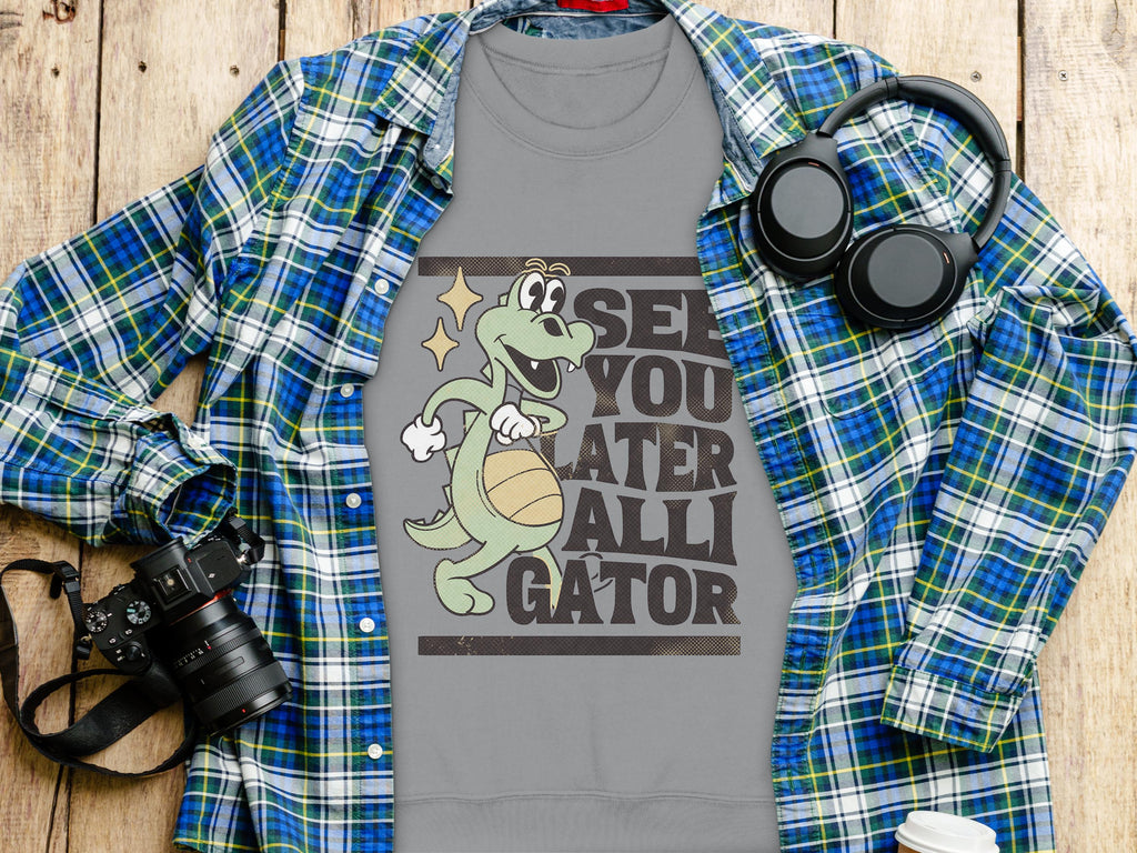 A Garment Graphics gray tee with a cartoon alligator holding a basketball and See You Later Alligator text is displayed on wood. Its surrounded by a blue plaid shirt, camera, headphones, and white travel mug.