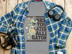 A Garment Graphics gray tee with a cartoon alligator holding a basketball and See You Later Alligator text is displayed on wood. Its surrounded by a blue plaid shirt, camera, headphones, and white travel mug.