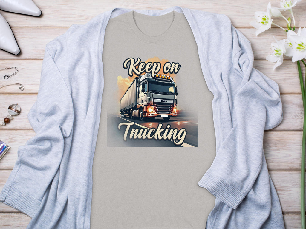 Garment Graphics T-shirt showcases a semi-truck graphic with Keep on Trucking text, perfect for truck enthusiasts. Its stylishly paired on a wooden surface with a light gray cardigan, jewelry, and a flower.