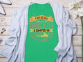 The Garment Graphics green T-shirt features the humorous quote: If hes got golf clubs in his truck or a camper in his driveway, I dont hire him. It makes an ideal gift and is styled on a flat surface with a gray cardigan, flowers, and accessories.