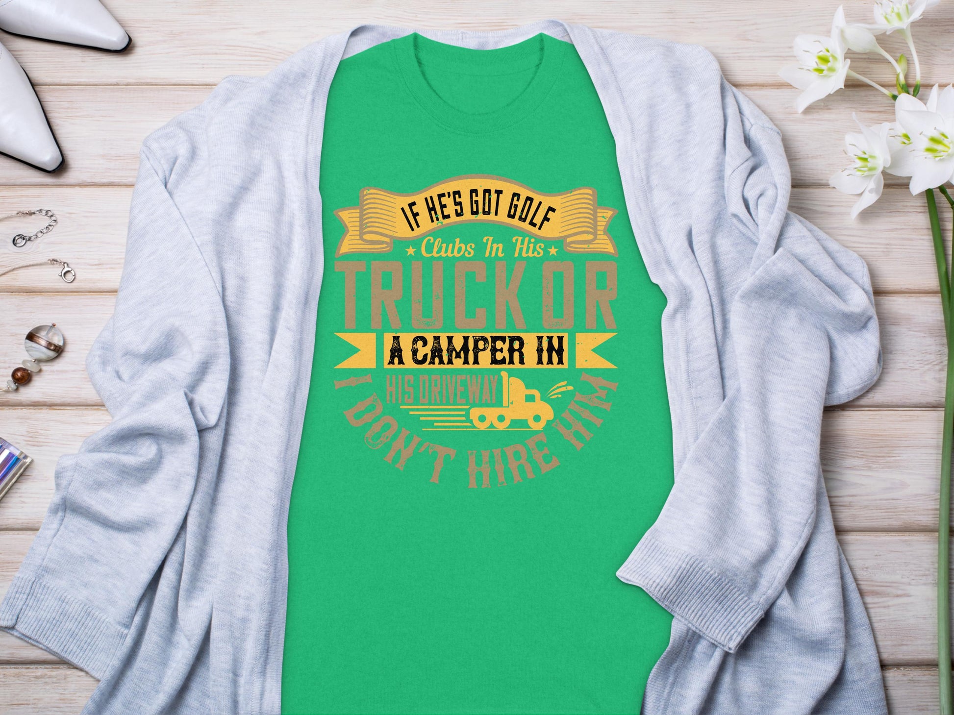 The Garment Graphics green T-shirt features the humorous quote: If hes got golf clubs in his truck or a camper in his driveway, I dont hire him. It makes an ideal gift and is styled on a flat surface with a gray cardigan, flowers, and accessories.
