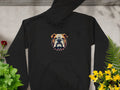 A Garment Graphics black hoodie features an embroidered stylized bulldog face with a spiked collar. It is displayed flat on concrete, surrounded by green, red, and yellow flowering plants.