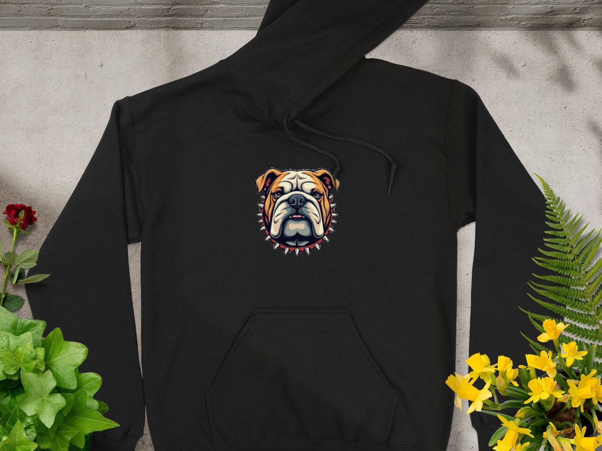 A Garment Graphics black hoodie features an embroidered stylized bulldog face with a spiked collar. It is displayed flat on concrete, surrounded by green, red, and yellow flowering plants.