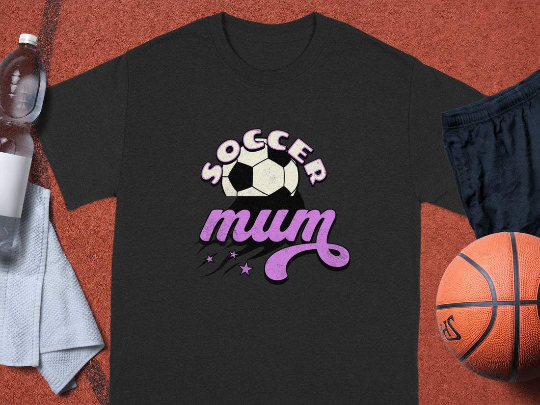 On a sandy path with grass behind, a person wears a classic fit T-shirt by Garment Graphics featuring SOCCER mum in black and pink letters, complete with a soccer ball and stars design.