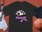 The Garment Graphics classic fit black T-shirt sports Soccer Mum in white and pink stylized text with a soccer ball graphic. Displayed on a reddish surface, it’s surrounded by a basketball, towel, navy shorts, and water bottle—ideal for fans of sporty style.