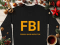 A Garment Graphics black sweatshirt with FBI and Female Boob Inspector text lies on a wooden table, surrounded by Christmas decorations. A cozy cup of coffee sits in the bottom right corner, enhancing the unisex appeal.