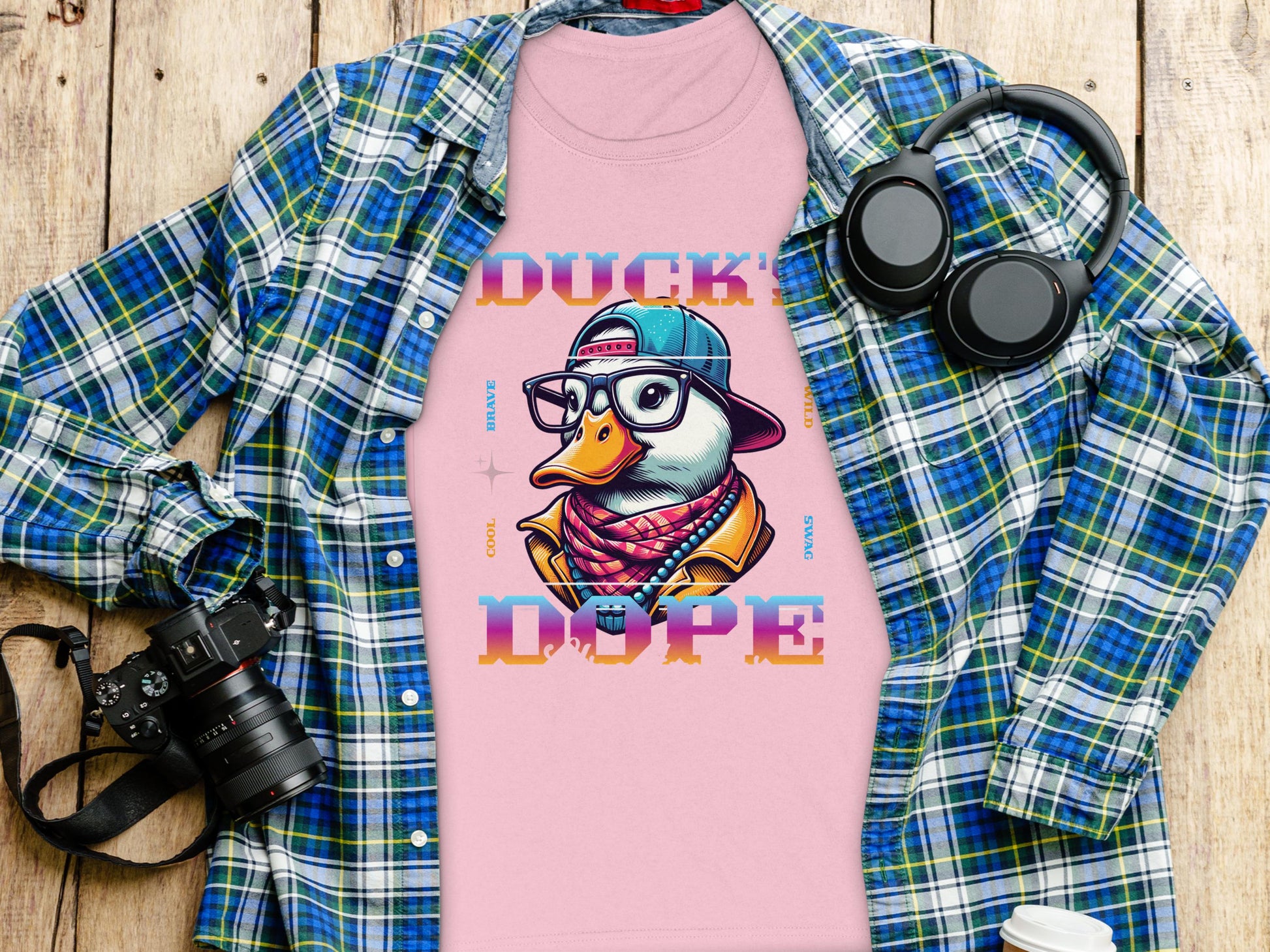 The pink t-shirt by Garment Graphics showcases a cartoon duck with glasses, cap, and scarf labeled DUCK DOPE. Exuding Cool Brave Wild Swag Duck vibes, its styled on a wooden surface under a blue plaid shirt with a camera, headphones, and coffee cup.