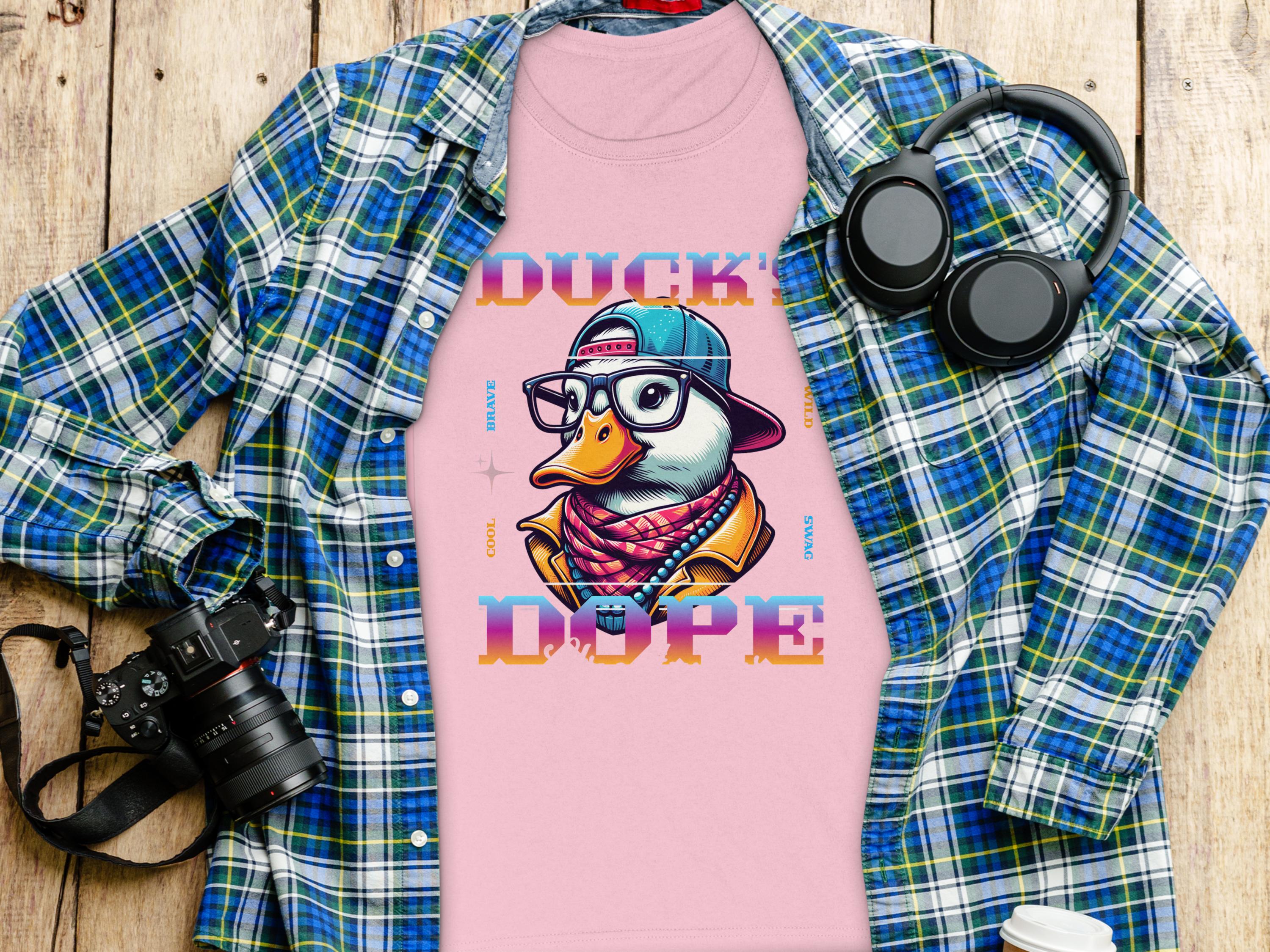 The pink t-shirt by Garment Graphics showcases a cartoon duck with glasses, cap, and scarf labeled DUCK DOPE. Exuding Cool Brave Wild Swag Duck vibes, its styled on a wooden surface under a blue plaid shirt with a camera, headphones, and coffee cup.