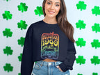 A woman with long brown hair smiles, exuding retro vibes in a Garment Graphics dark sweatshirt featuring a Woodstock 1969 design with a car and flowers. She pairs it with denim shorts against a backdrop of green shamrocks.