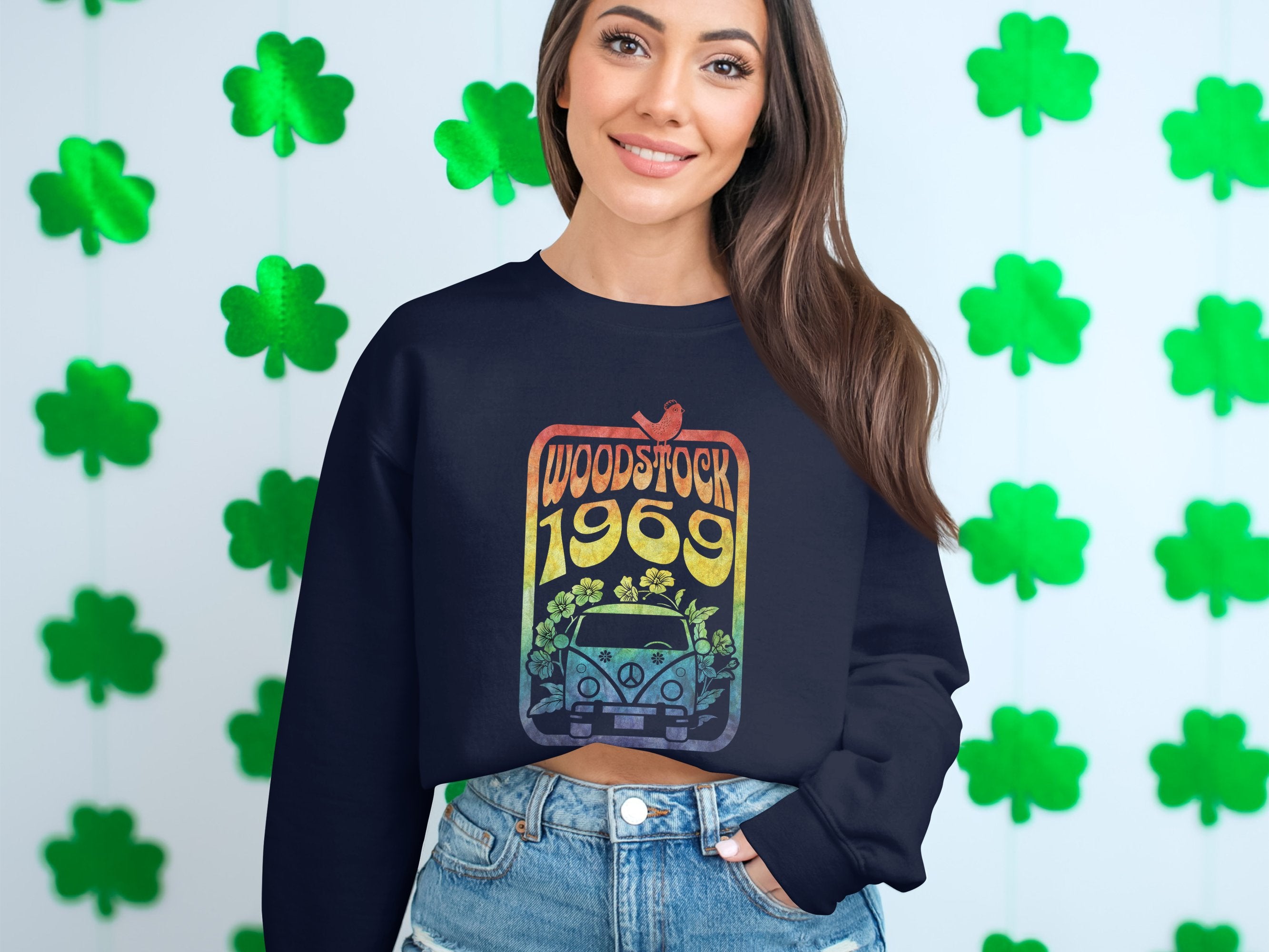 A woman with long brown hair smiles, exuding retro vibes in a Garment Graphics dark sweatshirt featuring a Woodstock 1969 design with a car and flowers. She pairs it with denim shorts against a backdrop of green shamrocks.