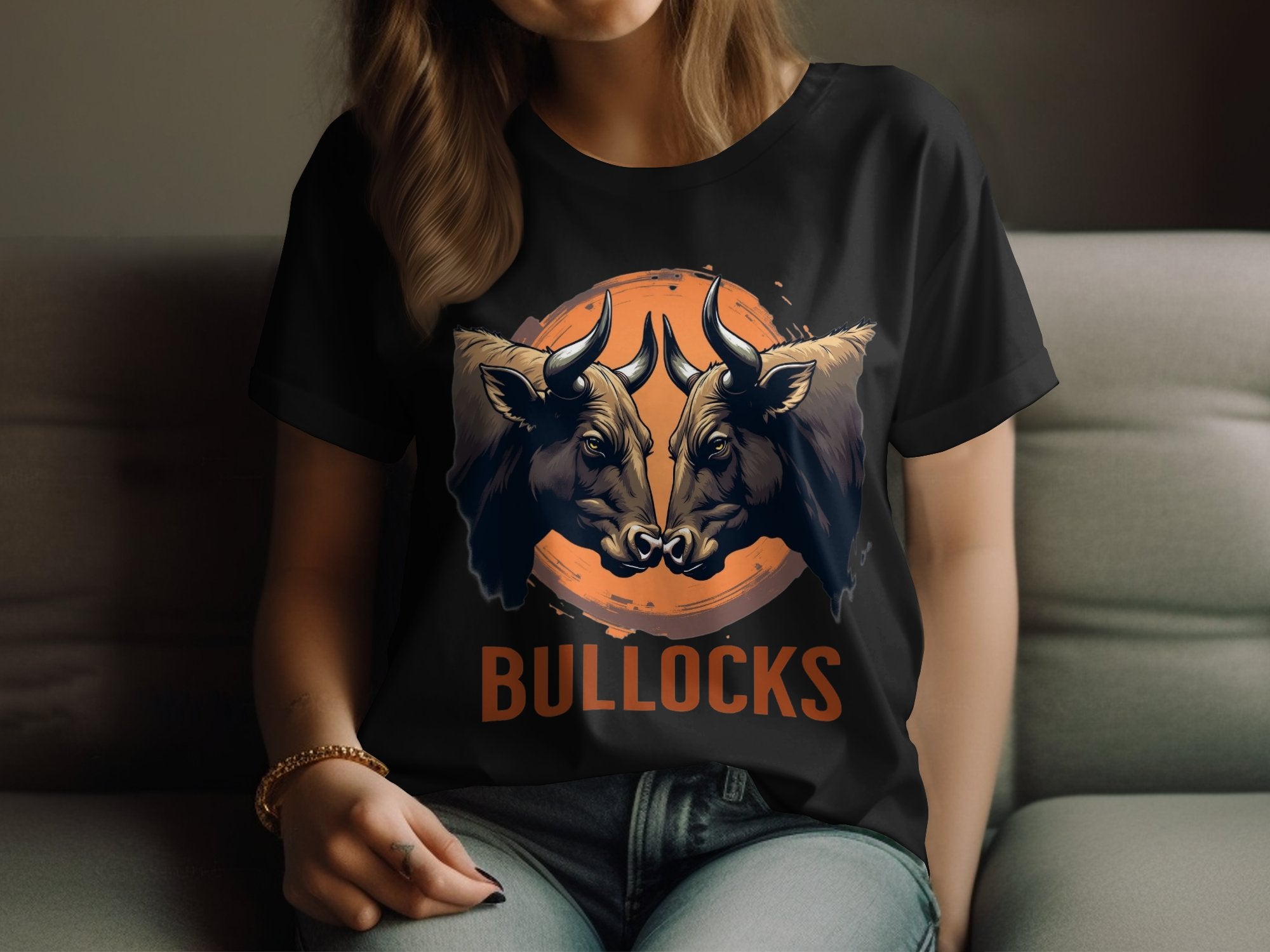 A person in casual wear sits on a couch, wearing a Garment Graphics black T-shirt with bold bull illustration graphics facing each other and BULLOCKS printed beneath. The head is partially out of frame.