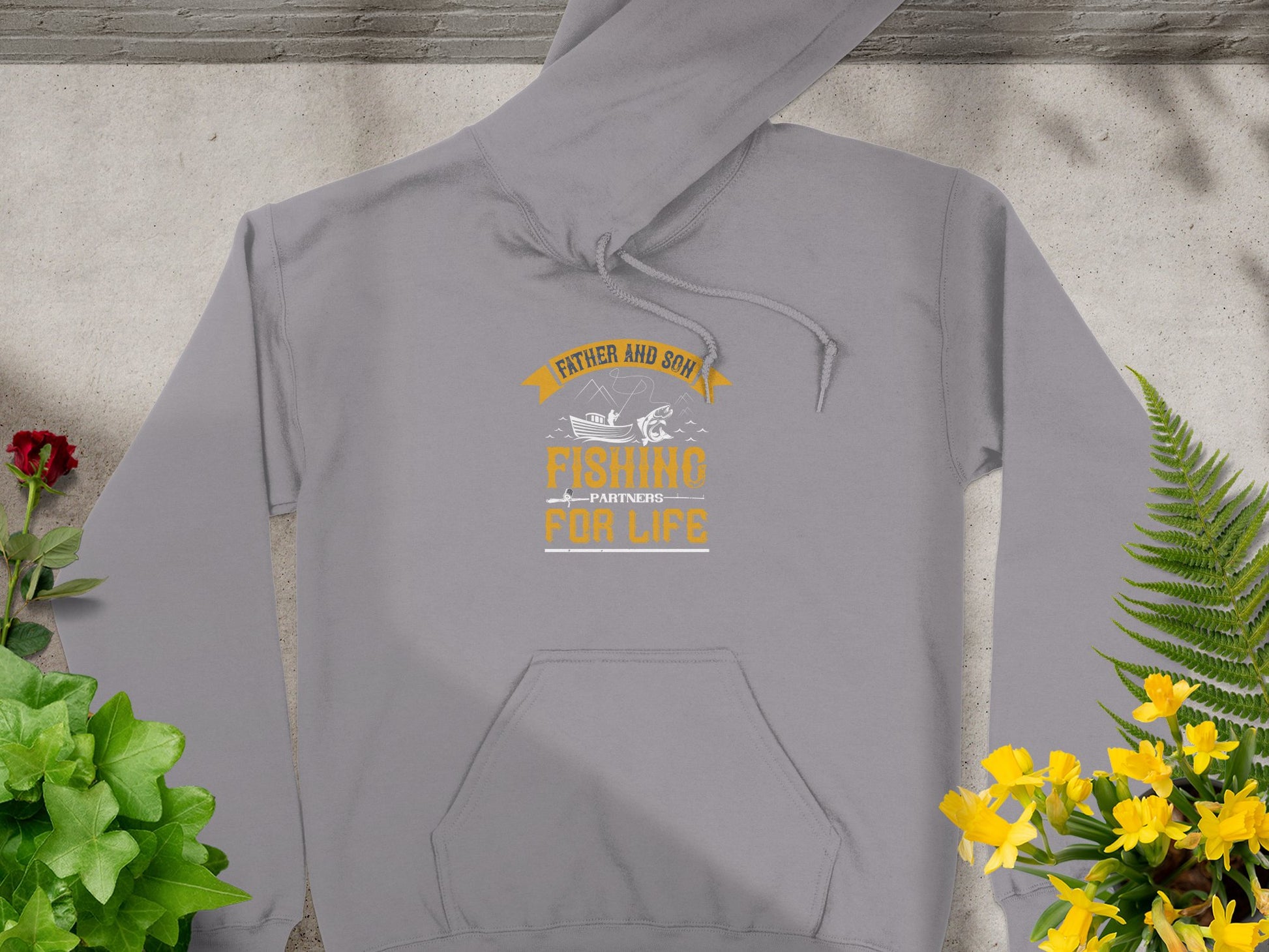 Garment Graphics gray fishing hoodie features the phrase Father and Son Fishing Partners for Life in orange and white lettering with a fish graphic. Perfect for fishing enthusiasts, its displayed on a light-colored background with flowers and greenery.