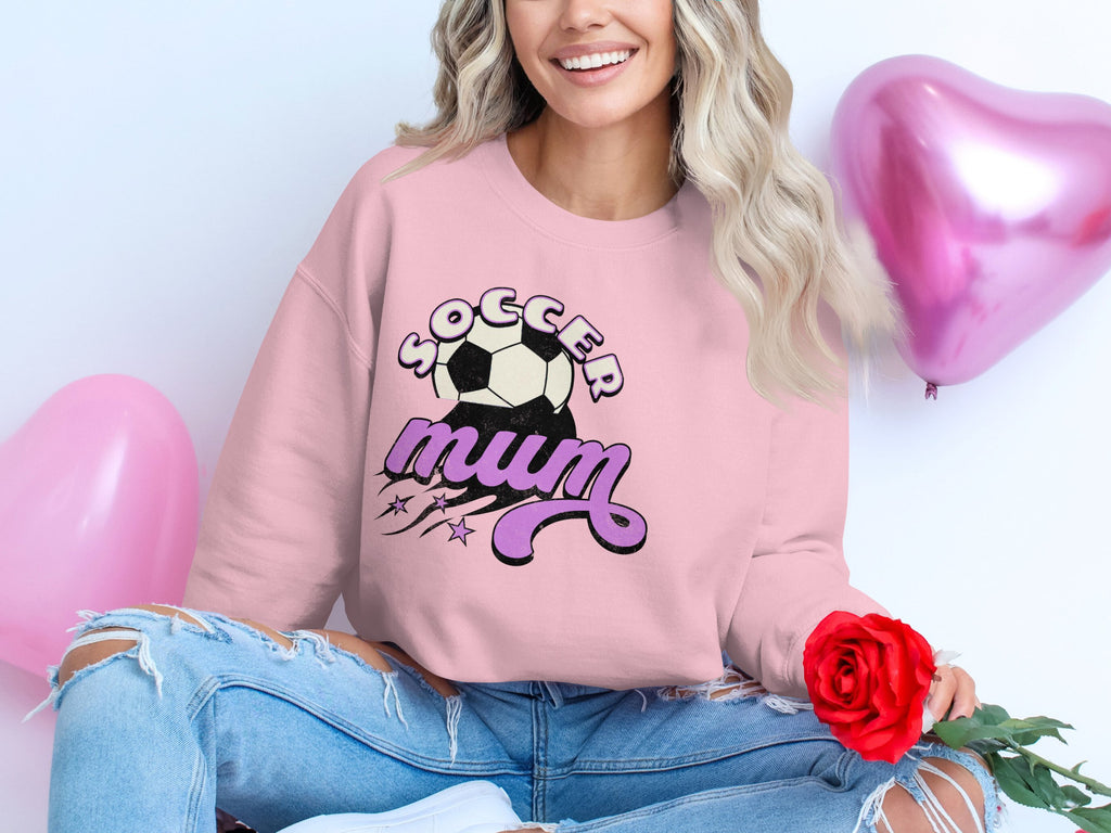 A seated person holds a red rose while wearing a Soccer Mum pink sweatshirt from Garment Graphics, paired with ripped jeans. The look is enhanced by pink heart-shaped balloons floating in the background.