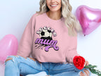 A seated person holds a red rose while wearing a Soccer Mum pink sweatshirt from Garment Graphics, paired with ripped jeans. The look is enhanced by pink heart-shaped balloons floating in the background.