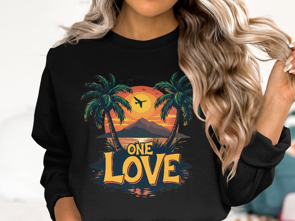 A person with wavy, long hair wears a black Garment Graphics sweatshirt featuring the vibrant One Love Sunset design, complete with palm trees silhouetted against a setting sun and birds.