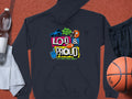 A black Garment Graphics hoodie displays vivid Loud & Proud, Lets Make Some Noise text with a megaphone, foam finger, and football graphics. It lies on an orange surface next to shorts, a basketball, towel, and water bottle.