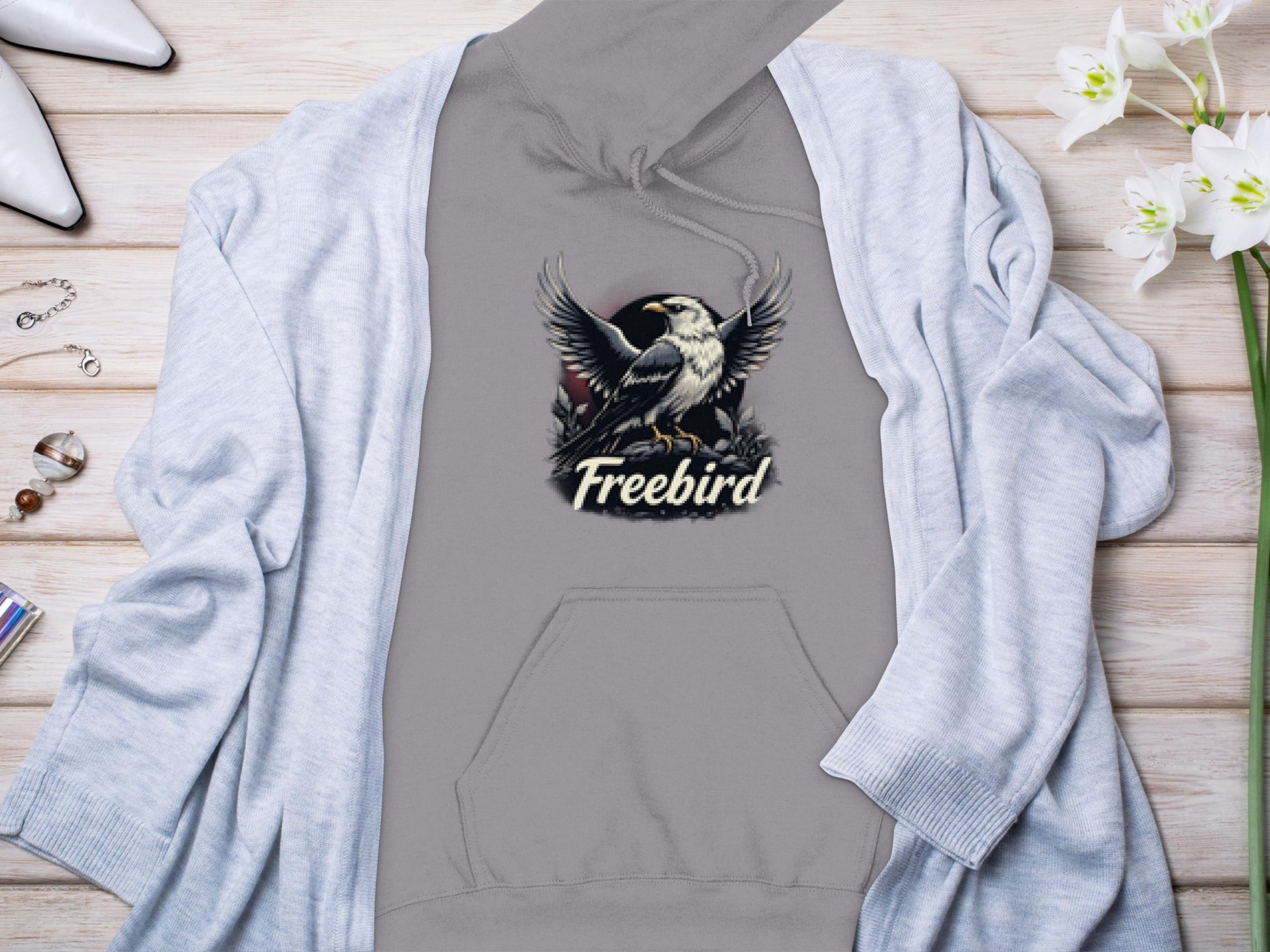 A Garment Graphics gray Freebird Graphic Pullover Hoodie, showcasing an eagle and the word Freebird, lies on a wooden surface, partly covered by a light gray cardigan. Surrounding it are flowers, jewelry, and white shoes that enhance its unique style.