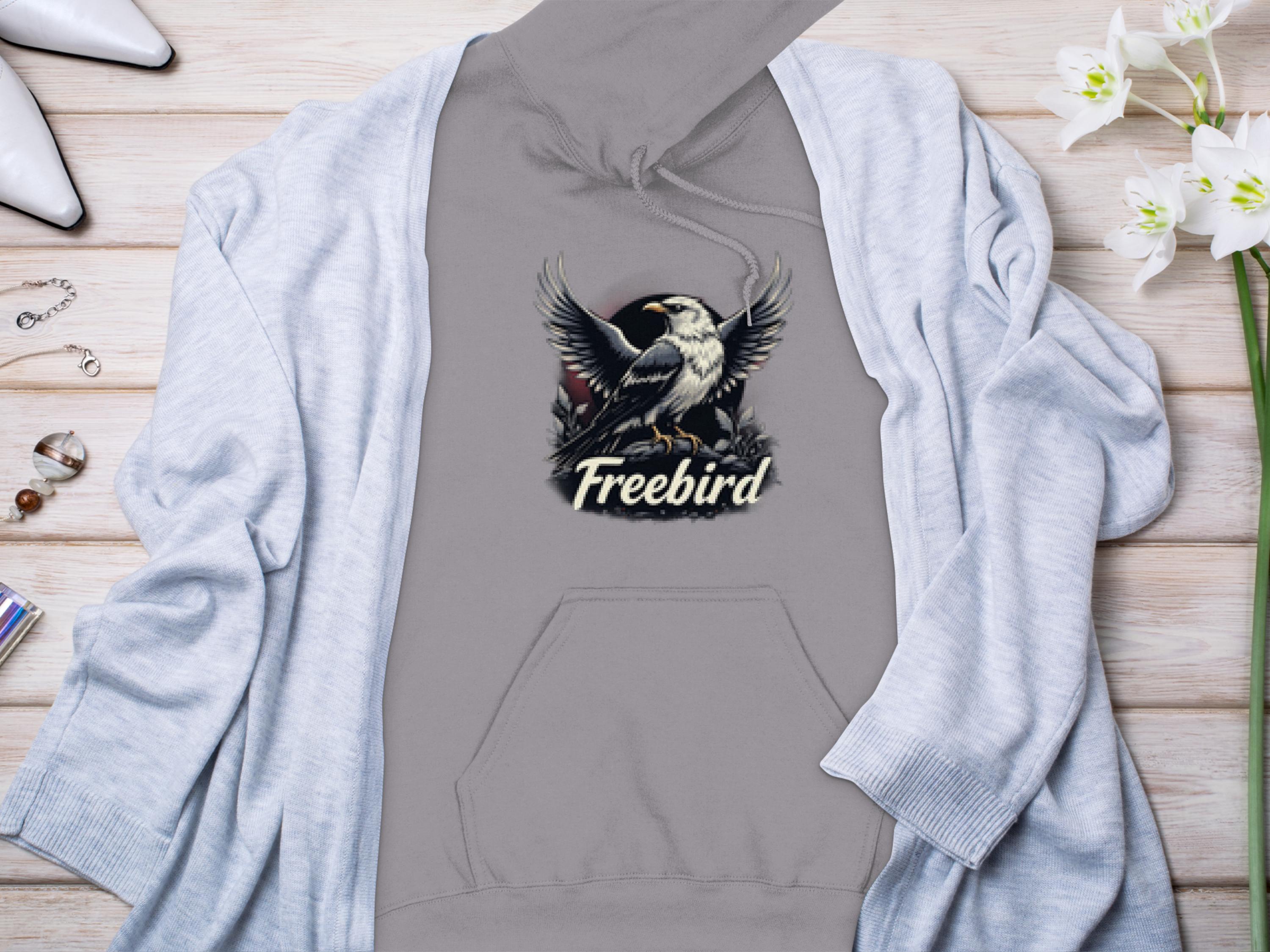 A Garment Graphics gray Freebird Graphic Pullover Hoodie, showcasing an eagle and the word Freebird, lies on a wooden surface, partly covered by a light gray cardigan. Surrounding it are flowers, jewelry, and white shoes that enhance its unique style.