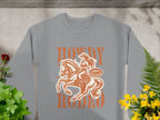 Garment Graphics presents a gray sweatshirt featuring a cowboy on horseback, ideal for rodeo fans. It boasts Howdy Rodeo and Not a Buckin Bronc Rider, surrounded by vivid red and yellow flowers with green leaves on a concrete backdrop. Capture the cowboy spirit in style.