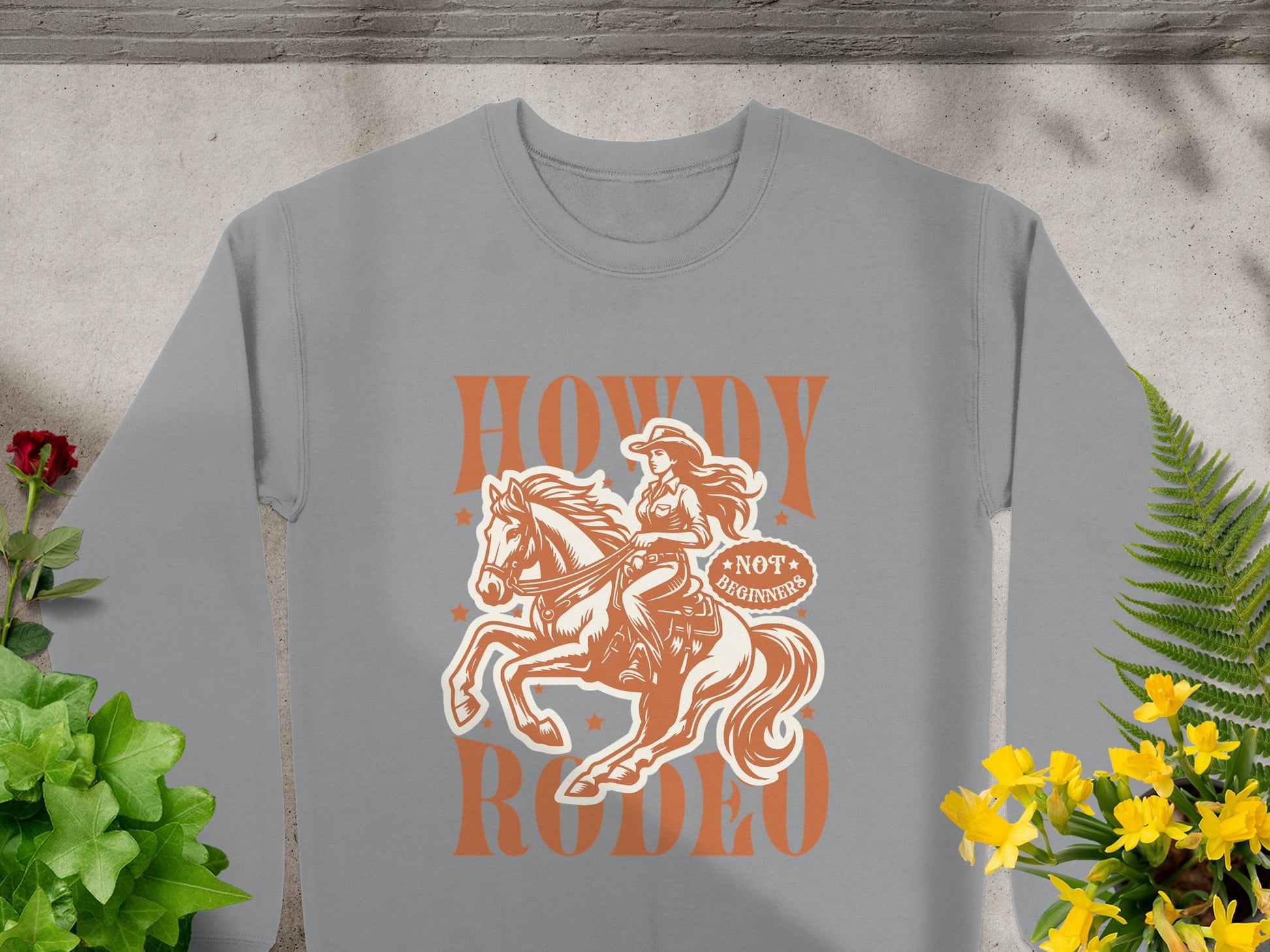 Garment Graphics presents a gray sweatshirt featuring a cowboy on horseback, ideal for rodeo fans. It boasts Howdy Rodeo and Not a Buckin Bronc Rider, surrounded by vivid red and yellow flowers with green leaves on a concrete backdrop. Capture the cowboy spirit in style.